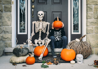 Full length of boy covering face with pumpkin while sitting by skeleton during Halloween