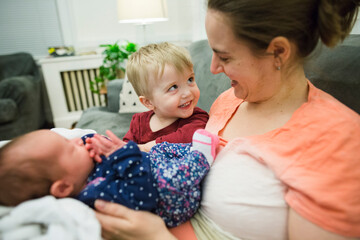 Happy blonde toddler boy looks up at mother holding newborn baby girl