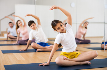 Girl sitting in lotus pose and doing exercises during group yoga training with her family in fitness studio.