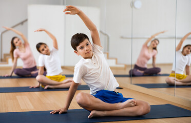 Smiling sporty tween boy doing stretching exercises while sitting in lotus position during training...