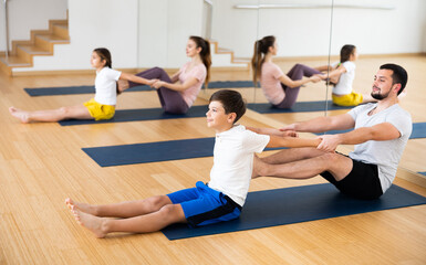 Fototapeta na wymiar Friendly sporty family practicing partner yoga in fitness studio. Smiling tween boy doing exercises on mat in pair with his father