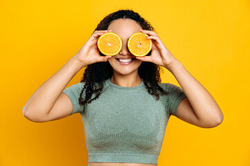 Healthy food and lifestyle, detox. Joyful lovely mixed race young curly haired woman, in sport outfits, standing on isolated orange background, holds two halves orange in her hands near eyes, smiling
