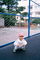 Little girl sits squatting on the asphalt near a metal fence. High quality photo