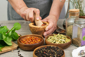 Woman's hands grind red pepper in a wooden mortar. Kitchen gray table with various spices.