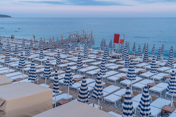 Rows of white beach beds with white and blue closed umbrellas lie on a French beach in Nice as...