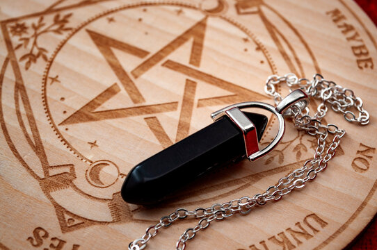 Dowsing pendulum with black gemstone next to hardwood divination chart on red velvet backgrounds concept for mystic magic spell, advanced esoteric symbols and secret occult philosophy