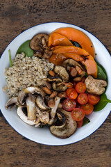 Top view of a nutritious salad with grilled vegetables. Vegetarian salad with grilled pumpkin, mushrooms, spinach, barley, almonds and cherry tomatoes, in a white bowl on the restaurant wooden table.