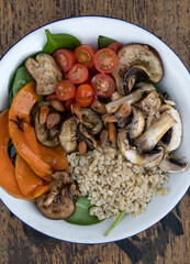 Top view of a nutritious salad with grilled vegetables. Vegetarian salad with grilled pumpkin, mushrooms, spinach, barley, almonds and cherry tomatoes, in a white bowl on the restaurant wooden table.