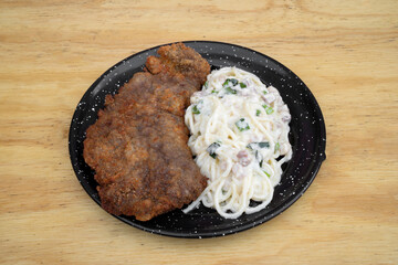 Pasta and meat. Top view of a milanesa, breaded steak, and spaghetti with a bacon and leek cream sauce, in a black dish on the restaurant wooden table.