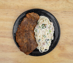 Pasta and meat. Top view of a milanesa, breaded steak, and spaghetti with a bacon and leek cream sauce, in a black dish on the restaurant wooden table. 