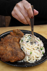 Eating at the restaurant. Closeup view of a woman's hand holding a fork, eating milanesa, breaded steak, and spaghetti with a bacon and leek creamy sauce.