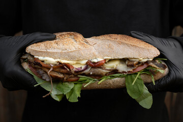 Chef with black gloves holding a big sandwich made with bread, grilled meat, arugula, cheese and onion. 