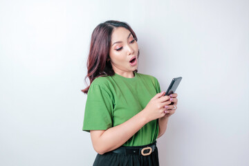 Fototapeta premium Surprised Asian woman wearing green t-shirt pointing at her smartphone, isolated by white background