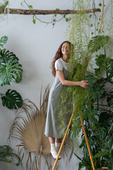 Young Spanish girl stands among green plants on stepladder and hugs flower in living room. Home comfort harmony. Symbol of purity, body care and nature harmony. Hobby, houseplant lovers concept © DimaBerlin