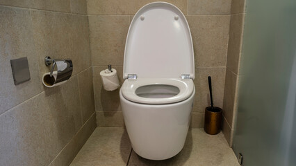 Restroom. White toilet in the center. Next - a cleaning brush, toilet paper rolls. The walls are made of beige tiles and frosted glass.