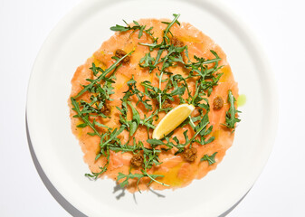 Salmon carpaccio with arugula on white background in summer day. Fish appetizer - salmon carpaccio in fine dining with shadows. Elegant carpaccio of raw salmon and sauce. Italian food. Aesthetic menu.