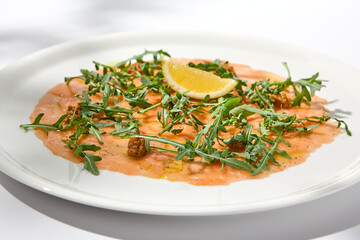 Salmon carpaccio with arugula on white background in summer day. Fish appetizer - salmon carpaccio in fine dining with shadows. Elegant carpaccio of raw salmon and sauce. Italian food. Aesthetic menu.
