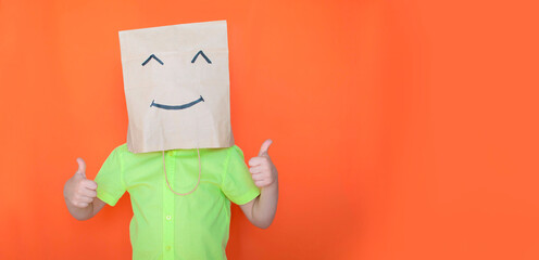 A child with a paper bag on his head in the form of a smiley face shows with two hands gesture of "OK", approval, sympathy. Double thumbs-up sign. to advertise a product or service. Space for copying