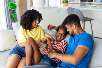 Happy multiethnic family sitting on sofa laughing together. Cheerful parents playing with their daughter at home. Father tickles his little girl while the mother is smiling.