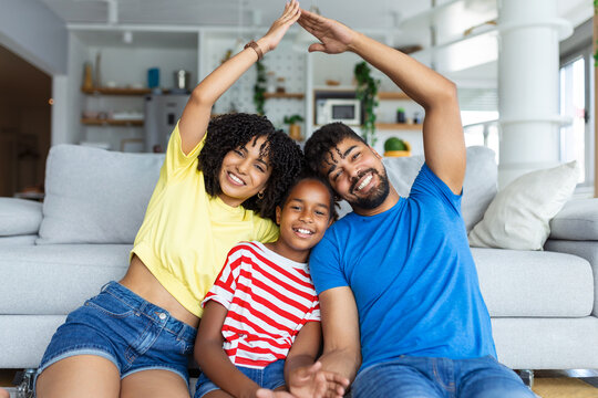 Family Care, Protection And Insurance Concept. Portrait of smiling African American parents making symbolic roof of hands above their happy daughet, sitting on the floor carpet in living room at home