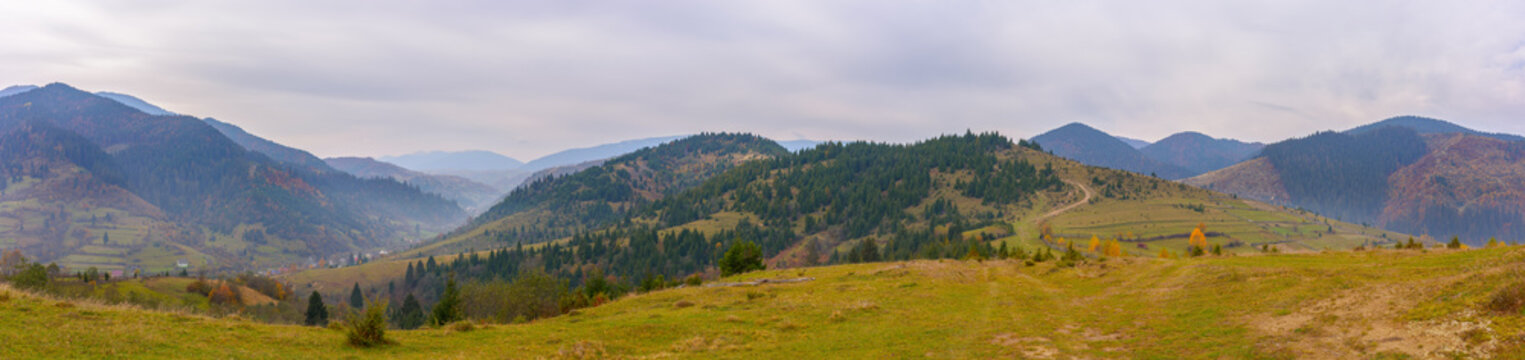 panorama of countryside landscape in autumn. grassy pasture meadows and forested hills. carpathian mountain ridge in the distance. village in the valley. overcast weather