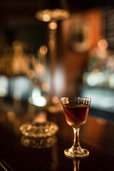 A glass with cold Manhattan cocktail garnished with orange zest peel on a wooden bar counter. Bokeh lights, selective focus, small depth of field