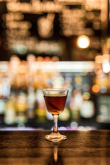 A glass with cold Manhattan cocktail garnished with orange zest peel on a wooden bar counter. Bokeh lights, selective focus, small depth of field