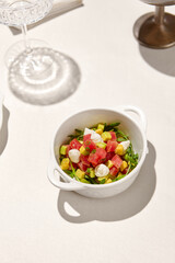 Tuna tartar in ceramic bowl on white table in sunny day. Tuna and avocado tartar with cream cheese in restaurant menu. Appetizer from tuna and avocado with shadow from wine glass.