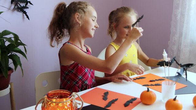 selective focus on two cute blonde schoolgirl sisters, they sit at a table and cut out Halloween symbols from paper, making decorations for the holiday.
