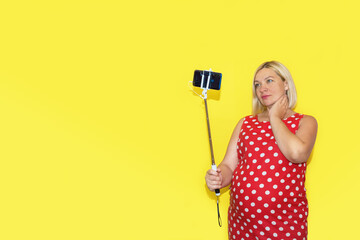 pregnant woman in a red polka dot dress takes a selfie on a yellow background with copy space