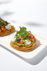Aesthetic composition with salmon bruschetta on white background with shadows from flowers. Italian bruschetta with salmon, avocado, cheese and olives on fine dining in summer. Elegant menu concept.