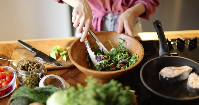Woman mixing salad and frying salmon steaks, cooking healthy food, close-up view with no face