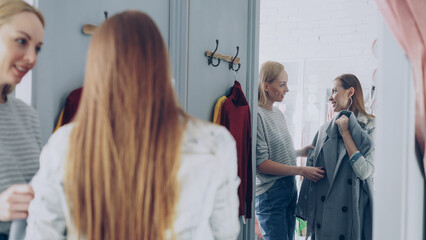 Pretty young woman is checking fashionable coat in fitting room with her friend helping her to...