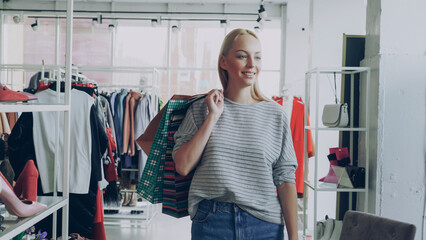 Dolly shot of blond girl walking between shelves and rails in large store. She is carrying lots of...