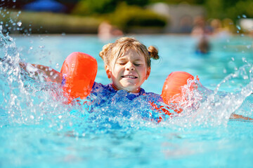 Little preschool girl with protective swimmies playing in outdoor swimming pool by sunset. Child learning to swim in outdoor pool, splashing with water, laughing and having fun. Family vacations.