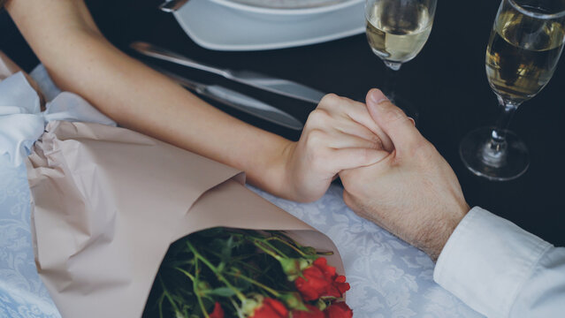 Close-up shot of male hand holding and squeezing manicured female hand on table with champagne glasses and flowers. Romantic relationship, love and fine dining concept.