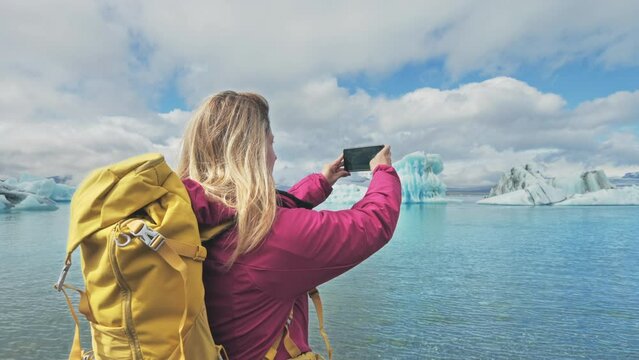 female hiker taking photo of  jokulsarlon iceberg glacier lake lagoon,woman traveler excursionist wearing backpack photographing icebergs floating on sea water using smartphone mobile