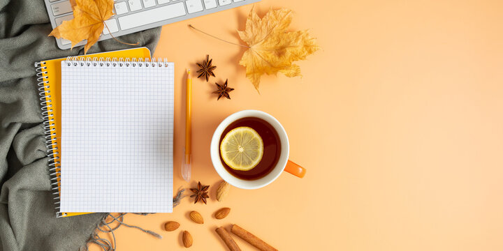 Autumn composition. Office desk with cup of hot tea with lemon, empty notebook, keyboard, scarf, dry leaves on isolated beige background. Autumn, fall business concept. Flat lay, top view, copy space