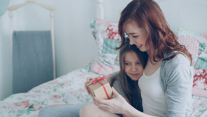 Obraz na płótnie Canvas Happy younf mother kissing her cute daughter presenting gift box on celebration sitting on bed in light cozy bedroom at home
