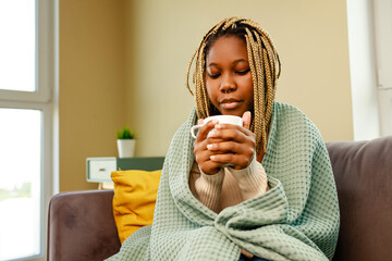 afro woman wrapped on blue blanket and drinking hot tea in living room