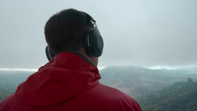 Handsome adult man listening to music through large headphones in the mountains on a stormy day looking at valley and misty volcano. Music-lover on a hiking trip wearing red jacket enjoying mount view
