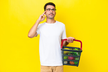 Young handsome man holding a shopping basket full of food over isolated yellow background having...