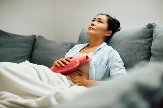 Young Asian woman holding hot water bottle on her stomach at home.
