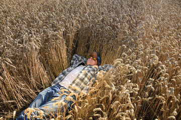 Young adult farmer relaxing at agricultural grain field at sunny day. Male worker wearing hat,...