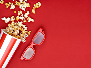 Popcorn spilled from a white and red striped bucket near 3D glasses on a red background. Top view, above, overhead. Space for text. Cinema, movies, movie night and entertainment concept.