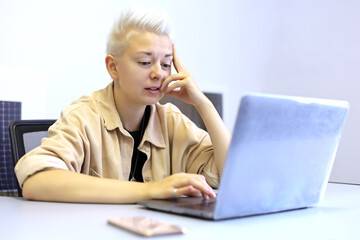 Girl with thoughtful expression and short blonde hair sitting at office table with laptop. Tomboy lifestyle, concept of inspiration at work and creativity