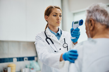 Female doctor measuring temperature of senior patient with infrared thermometer at medical clinic.