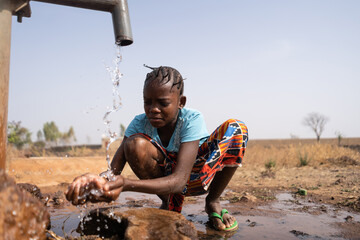 A young African girl casually crouches in front of a water pump and washes roughly, which is...