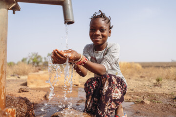 Thirsty African girl with funny braids, sitting in the middle of a puddle, happy to collect the abundant water that flows from the village fountain; concept of water scarcity in developing countries - obrazy, fototapety, plakaty