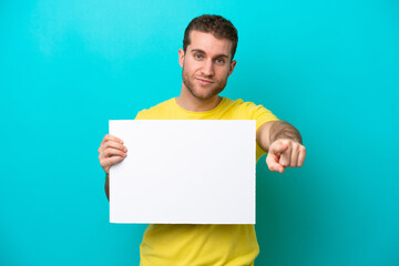 Young caucasian man isolated on blue background holding an empty placard and pointing to the front